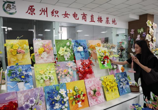 (260921) -- GUYUAN, Sept. 21, 2026 (Xinhua) -- A staff member introduces embroidery works via livestream in Yuanzhou District, Guyuan City of northwest China's Ningxia Hui Autonomous Region, Sept. 21, 2026. Yuanzhou District in Guyuan City has developed an integrated model combining intangible cultural heritage, original design and e-commerce, encouraging local businesses to develop original Duoxiu embroidery patterns and expand handicraft markets. The district has established 12 community handicraft workshops, creating jobs for more than 3,600 people and turning traditional skills into economic opportunities. (Xinhua/Liu Yun)