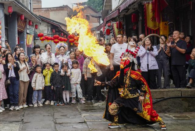(260923) -- CHONGQING, Sept. 23, 2026 (Xinhua) -- Visitors enjoy a Sichuan Opera fire-breathing performance at the ancient town of Zouma in Chongqing, southwest China, Sept. 23, 2026. As the Mid-Autumn Festival and National Day holiday period approaches, the ancient town of Zouma has launched a series of folk cultural activities, including lion dance performances, an intangible cultural heritage market, a Mid-Autumn lantern riddle fair, Hanfu experiences, and Sichuan Opera face-changing performances. Integrating the historical heritage of the town's ancient post road with contemporary folk culture experiences, these activities have attracted numerous local residents and tourists. (Xinhua/Liu Chan)