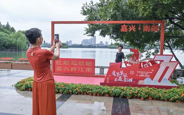 (261001) -- BEIJING, Oct. 1, 2026 (Xinhua) -- A tourist poses for a photo with an installation themed on National Day celebrations at the Nanhu Lake scenic area in Jiaxing, east China's Zhejiang Province, Oct. 1, 2026. People celebrated the 77th anniversary of the founding of the People's Republic of China in various ways on Thursday. (Xinhua/Xu Yu)