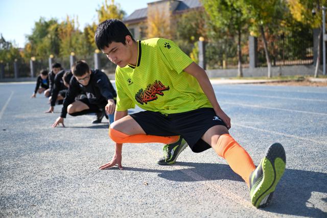(261015) -- URUMQI, Oct. 15, 2026 (Xinhua) -- Players warm up before a daily training at Xinjiang Special Education Vocational Secondary School in Changji Hui Autonomous Prefecture, northwest China's Xinjiang Uygur Autonomous Region, Oct. 14, 2026. Xinjiang's first blind football team is known as the 'Wei Wei Wei' team. It takes its name from the call players shout to locate teammates and avoid collisions. This call is the communication code of this special sport.
In blind football, players wear light-blocking blindfolds throughout the match. They track the ball by the bell inside it and keep calling 'wei, wei, wei' to gauge teammates' positions, enabling them to dribble, pass and shoot without sight. In training, they run, collide, feel the boundary boards, and build muscle memory through countless falls and getting back up. For visually impaired teenagers who rarely dare to sprint at full speed in daily life, this enclosed pitch is a place where they can run freely.
'We cannot see the world, but we strive to make the world see us.' In the darkness, guided by their calls and the sound of the ball, these young athletes break through barriers and shine brightly on the green pitch. (Xinhua/Ding Lei)