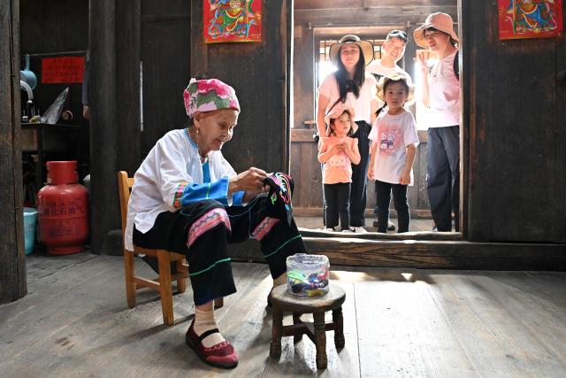 (261016) -- LONGSHENG, Oct. 16, 2026 (Xinhua) -- Tourists look on as Hou Yujin (front) works on an embroidery work in Longji Village of Longsheng County, south China's Guangxi Zhuang Autonomous Region, Oct. 15, 2026. Longji Village, home mainly to the Zhuang ethnic group, features traditional wooden houses nestled among terraced fields and retains its distinctive ethnic cultural heritage.
   Hou Yujin, an elderly local woman who lives in one of the century-old stilted buildings of Zhuang ethnic group in the village, has volunteered to offer free guided tours to tourists. Despite never having attended a single day of school, Hou taught herself Mandarin through great efforts to share with visitors the history of her house and the dramatic changes of the village. (Xinhua/Lu Boan)