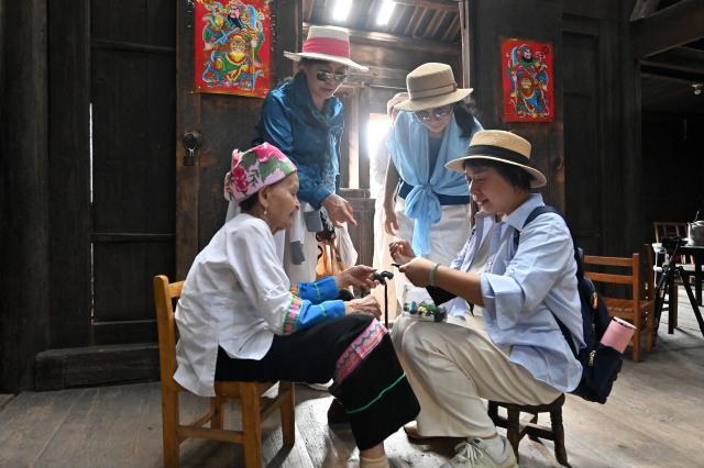 (261016) -- LONGSHENG, Oct. 16, 2026 (Xinhua) -- Hou Yujin (L, front) communicates with tourists about embroidery techniques in Longji Village of Longsheng County, south China's Guangxi Zhuang Autonomous Region, Oct. 15, 2026. Longji Village, home mainly to the Zhuang ethnic group, features traditional wooden houses nestled among terraced fields and retains its distinctive ethnic cultural heritage.
   Hou Yujin, an elderly local woman who lives in one of the century-old stilted buildings of Zhuang ethnic group in the village, has volunteered to offer free guided tours to tourists. Despite never having attended a single day of school, Hou taught herself Mandarin through great efforts to share with visitors the history of her house and the dramatic changes of the village. (Xinhua/Lu Boan)