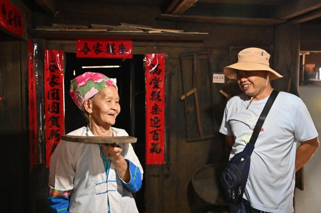 (261016) -- LONGSHENG, Oct. 16, 2026 (Xinhua) -- Hou Yujin (L) introduces an old item to a tourist at her stilted house in Longji Village of Longsheng County, south China's Guangxi Zhuang Autonomous Region, Oct. 15, 2026. Longji Village, home mainly to the Zhuang ethnic group, features traditional wooden houses nestled among terraced fields and retains its distinctive ethnic cultural heritage.
   Hou Yujin, an elderly local woman who lives in one of the century-old stilted buildings of Zhuang ethnic group in the village, has volunteered to offer free guided tours to tourists. Despite never having attended a single day of school, Hou taught herself Mandarin through great efforts to share with visitors the history of her house and the dramatic changes of the village. (Xinhua/Lu Boan)