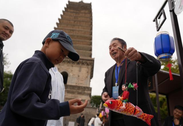 (261017) -- SHENYANG, Oct. 17, 2026 (Xinhua) -- Sun Yu'en (R) displays his Chaoyang red clay figure work for a child at a market for intangible cultural heritage in Chaoyang, northeast China's Liaoning Province, Oct. 16, 2026. Chaoyang red clay figure is a provincial-level intangible cultural heritage in Liaoning Province, northeast China. 
  Sun Yu'en, an inheritor of this craft, has loved the art since childhood. In 1998, he devoted himself to the craft and learned the core skills from his teacher. Sun's artworks emphasize the expressions and details of the figures, embodying rustic charm. He has also created a series of moving clay figures, which are popular among young people. (Xinhua/Li Gang)