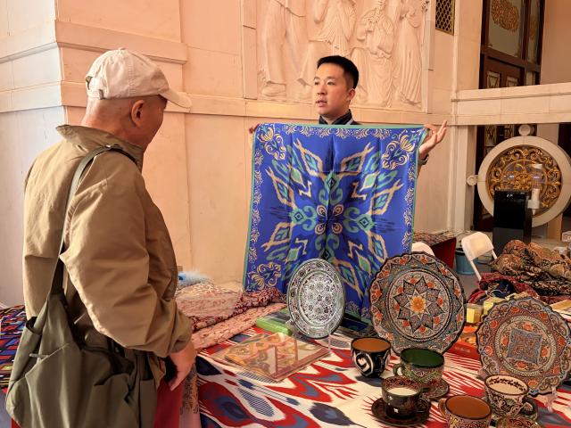 (261021) -- BEIJING, Oct. 21, 2026 (Xinhua) -- Gong Yue, a young entrepreneur from Korla, northwest China's Xinjiang Uygur Autonomous Region, shows a scarf he designed with traditional elements of Xinjiang to a visitor at an exhibition in Beijing, capital of China, Oct. 16, 2026. TO GO WITH "Across China: Young entrepreneurs give Xinjiang's cultural heritage modern makeover" (Xinhua/Ji Xiaodong)