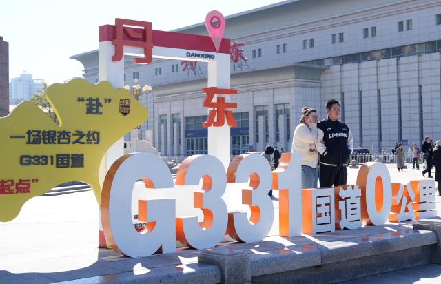 (261023) -- DANDONG, Oct. 23, 2026 (Xinhua) -- Tourists pose for a photo with a starting-point marker of the G331 national highway in Dandong, northeast China's Liaoning Province, Oct. 22, 2026. Autumn scenery and cultural attractions are drawing tourists to Dandong, boosting local tourism industry. (Xinhua/Li Gang)