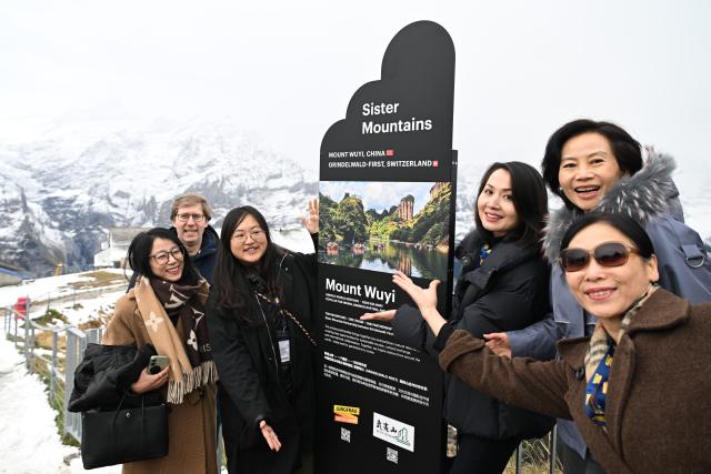 (261025) -- GENEVA, Oct. 25, 2026 (Xinhua) -- Guests pose for group photos with a display board marking the sister-peaks partnership between Yunv Peak, a landmark of China's Wuyi Mountain, and Switzerland's Grindelwald-First mountain in the canton of Bern, Switzerland, Oct. 23, 2026. TO GO WITH "Feature: Taste of China's Wuyi Mountain at Switzerland's Grindelwald-First" (Xinhua/Lian Yi)