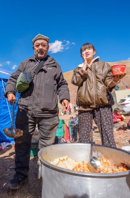 (261027) -- BAQEN, Oct. 27, 2026 (Xinhua) -- Alimjan Haliq (L) and Zhe Jiaxin serve pilaf for residents at a temporary shelter in Maru Township of Baqen County, Nagqu City, southwest China's Xizang Autonomous Region, Oct. 26, 2026.
  In the wake of the earthquake that struck Baqen County on Oct. 22, several volunteers rushed to the quake-affected zone to serve hot meals, sending warmth to those affected who are facing high-altitude cold in late October.
   Among them was Alimjan Haliq, a volunteer from northwest China's Xinjiang. Knowing how difficult it is to cook at high altitude, he brought two pressure cookers with him all the way to the quake zone. Zhe Jiaxin, a young woman from Xinjiang, spent nearly 48 hours on a bumpy journey through multiple cities before finally reaching the epicenter. Abdu, who runs a Xinjiang restaurant in Lhasa, quickly rallied his friends, gathered supplies, and rushed to the disaster area. Their paths were different, but they converged on the snow-covered plateau for the same purpose.
   "There is strength in numbers," Alimjan Haliq said, voicing the thoughts of every volunteer. Upon arriving in the quake zone, the rows of tidy tents and prefab houses, ample supplies, and locals helping one another deeply moved the volunteers. Zhe Jiaxin said with emotion, "I want to do all I can to help the children here feel more comfortable."
   The unceasing cook fires and hot food from the volunteers did more than fill empty stomachs -- they let the affected people feel be cared and supported. (Xinhua/Tenzin Nyida)