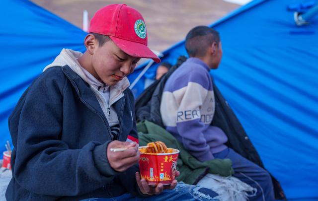 (261027) -- BAQEN, Oct. 27, 2026 (Xinhua) -- A child eats stir-fried rice noodles at a temporary shelter in Maru Township of Baqen County, Nagqu City, southwest China's Xizang Autonomous Region, Oct. 26, 2026.
  In the wake of the earthquake that struck Baqen County on Oct. 22, several volunteers rushed to the quake-affected zone to serve hot meals, sending warmth to those affected who are facing high-altitude cold in late October.
   Among them was Alimjan Haliq, a volunteer from northwest China's Xinjiang. Knowing how difficult it is to cook at high altitude, he brought two pressure cookers with him all the way to the quake zone. Zhe Jiaxin, a young woman from Xinjiang, spent nearly 48 hours on a bumpy journey through multiple cities before finally reaching the epicenter. Abdu, who runs a Xinjiang restaurant in Lhasa, quickly rallied his friends, gathered supplies, and rushed to the disaster area. Their paths were different, but they converged on the snow-covered plateau for the same purpose.
   "There is strength in numbers," Alimjan Haliq said, voicing the thoughts of every volunteer. Upon arriving in the quake zone, the rows of tidy tents and prefab houses, ample supplies, and locals helping one another deeply moved the volunteers. Zhe Jiaxin said with emotion, "I want to do all I can to help the children here feel more comfortable."
   The unceasing cook fires and hot food from the volunteers did more than fill empty stomachs -- they let the affected people feel be cared and supported. (Xinhua/Tenzin Nyida)