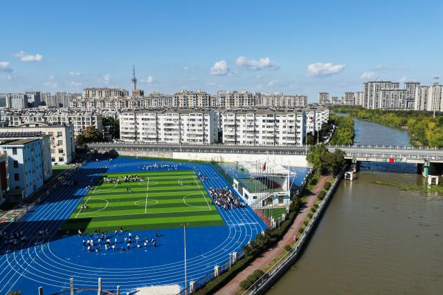 (261028) -- RUDONG, Oct. 28, 2026 (Xinhua) -- An aerial drone photo taken on Oct. 27, 2026 shows students exercising on a playground near the Jueju River in Rudong County of Nantong City, east China's Jiangsu Province. In recent years, Rudong County in Nantong City has made efforts to improve its aquatic ecological environment. Water environment of the three rivers, namely the Taiyun River, the Jueju River and the Juekan River, which run through the county, has undergone sound remediation. Along these waterways, the Heming Park and the bank areas of tributaries have all formed a picturesque urban landscape for people to enjoy their leisure time. (Xinhua/Ji Chunpeng)